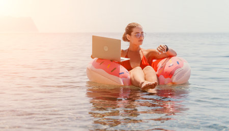 Woman freelancer works on laptop swimming in sea on pink inflatable ring. Happy tourist floating on...