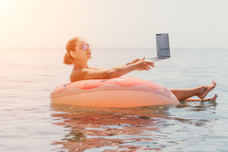 Woman freelancer works on laptop swimming in sea on pink inflatable ring. Happy tourist floating on...