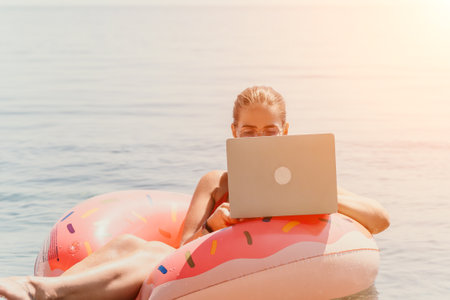 Woman freelancer works on laptop swimming in sea on pink inflatable ring. Pretty lady typing on computer while floating in the sea on inflatable donut at sunset. Freelance, remote work on vacationの写真素材