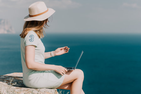Digital nomad, woman in the hat, a business woman with a laptop sits on the rocks by the sea during...