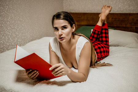 Young brunette woman works and studies from home on a laptop, while lying on her bed in red...
