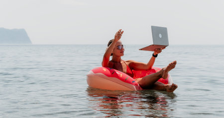 Woman freelancer works on laptop swimming in sea on pink inflatable ring. Happy tourist floating on inflatable donut and working on laptop computer in calm ocean. Freelance, remote working anywhereの写真素材