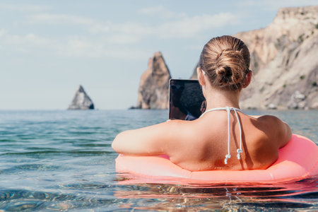 Woman freelancer works on laptop swimming in sea on pink inflatable ring. Happy tourist in...