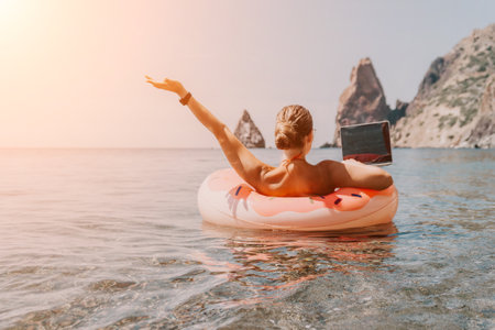 Woman freelancer works on laptop swimming in sea on pink inflatable ring. Happy tourist floating on inflatable donut and working on laptop computer in calm ocean. Freelance, remote working anywhereの写真素材