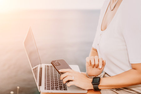 Digital nomad, Business woman working on laptop by the sea. Pretty lady typing on computer by the sea at sunset, makes a business transaction online from a distance. Freelance, remote work on vacationの写真素材