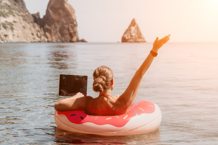Woman freelancer works on laptop swimming in sea on pink inflatable ring. Happy tourist floating on inflatable donut and working on laptop computer in calm ocean. Freelance, remote working anywhereの写真素材
