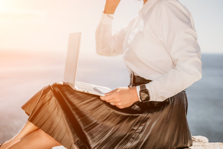 Digital nomad, Business woman working on laptop by the sea. Pretty lady typing on computer by the sea at sunset, makes a business transaction online from a distance. Freelance, remote work on vacationの写真素材