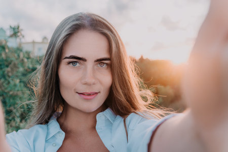 Amber Golden Hour Close up of a woman bathed in golden hour light making selfie on sunset in summer park. Happy tourist enjoy taking selfie photo outdoors for memoriesの写真素材