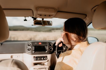 A woman in a yellow shirt is driving a car. She is wearing glasses and has her hands on the steering...