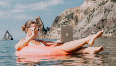 Woman freelancer works on laptop swimming in sea on pink inflatable ring. Happy tourist in...