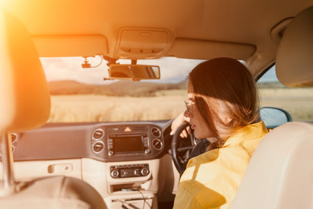 A woman in a yellow shirt is driving a car. She is wearing glasses and has her hands on the steering...