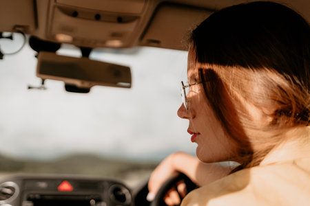 A woman in a yellow shirt is driving a car. She is wearing glasses and has her hands on the steering...