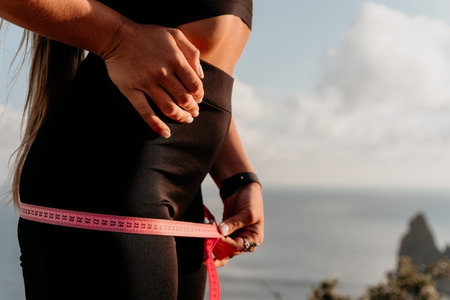 A woman is holding a pink tape measure around her hips. She is wearing black pants and a black top.