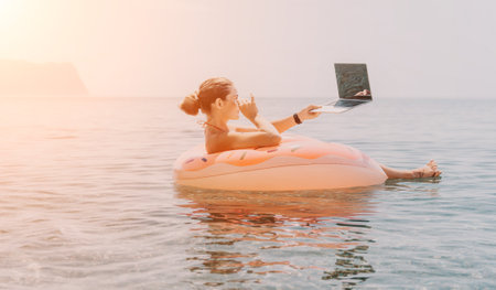 Woman freelancer works on laptop swimming in sea on pink inflatable ring. Happy tourist floating on inflatable donut and working on laptop computer in calm ocean. Freelance, remote working anywhereの写真素材