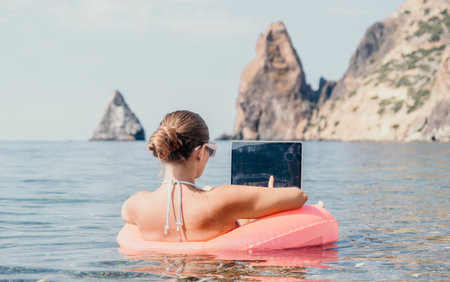 Woman freelancer works on laptop swimming in sea on pink inflatable ring. Happy tourist in...