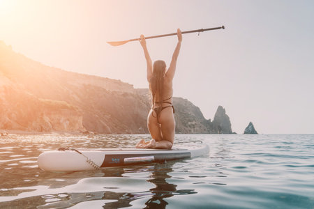 Woman Paddleboarding Ocean Coastline - A woman in a swimsuit kneels on a paddleboard in the ocean, raising her arms in the air. The coastline with cliffs is visible in the distance.の写真素材