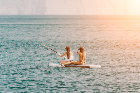 Two women are on a surfboard in the ocean. One of them is wearing a white shirt and the other is wearing a black bikini.の写真素材