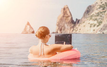 Woman freelancer works on laptop swimming in sea on pink inflatable ring. Happy tourist in sunglasses floating on inflatable donut and working on laptop computer in calm ocean. Remote working anywhereの写真素材