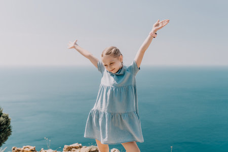 A young girl is standing on a beach, wearing a blue dress and holding her arms up in the air. Concept of joy and freedom, as the girl appears to be enjoying her time by the ocean.の写真素材