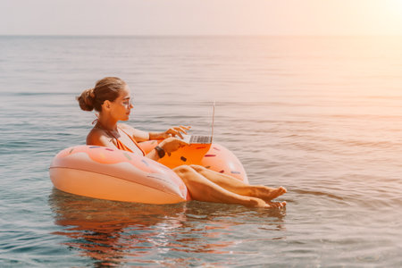 Woman freelancer works on laptop swimming in sea on pink inflatable ring. Happy tourist floating on inflatable donut and working on laptop computer in calm ocean. Freelance, remote working anywhereの写真素材