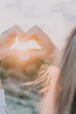 A womans hand is holding a heart shape, with the sun shining on it. Concept of love and warmth, as...