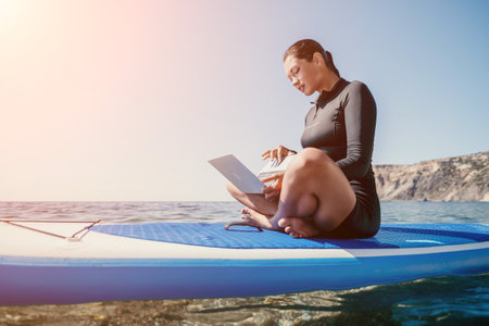 Woman sea laptop. Digital nomad, freelancer with laptop working on sup board at calm sea beach. Happy smiling girl relieves stress from work. Freelance, digital nomad, travel and holidays conceptの写真素材