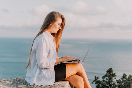 Woman Laptop Ocean - Working Remotely on Cliff Overlooking Sea at Sunsetの写真素材