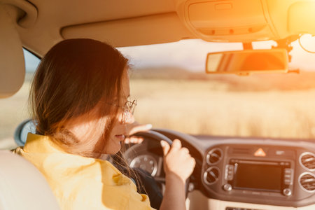 A woman in a yellow shirt is driving a car. She is wearing glasses and has her hands on the steering wheelの写真素材