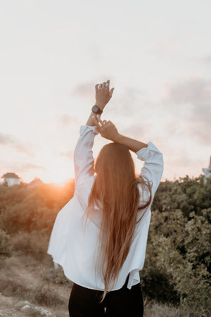Woman Sunset Outdoors Reaching for Sky - A woman with long hair stands in a field, with her arms raised towards the sunset.の写真素材