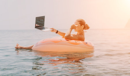 Woman Laptop Beach - A woman is sitting on an inflatable raft in the ocean using her laptop computer while enjoying a beach day.の写真素材