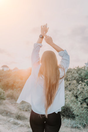 A woman raises her arms in the air with the sun setting behind her on a mountain.の写真素材