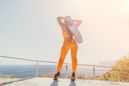 Woman in Orange Outfit with Yoga Mat, Standing on a Cliff overlooking the Seaの写真素材