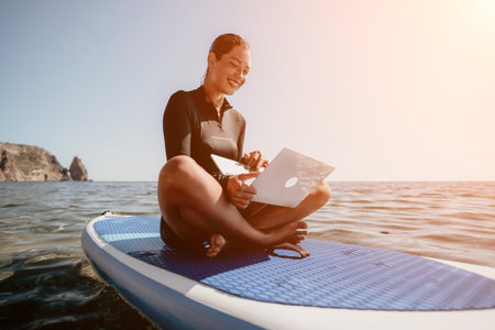 Woman Laptop Paddleboard - Smiling woman sits on paddleboard on blue ocean water, working on a laptop computer.の写真素材