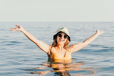 Woman Beach Summer - Happy woman in yellow swimsuit and bucket hat stands in ocean with arms raised.の写真素材