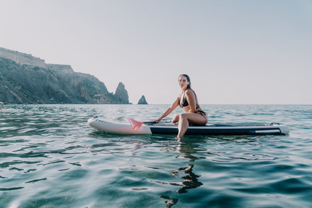 Woman Paddleboarding Ocean Coastline Summer - A woman in a bikini sits on a paddleboard in the ocean, with a mountainous coastline in the background. The sun is shining and the water is calm. This is a popular summer activity for people of all ages.の写真素材