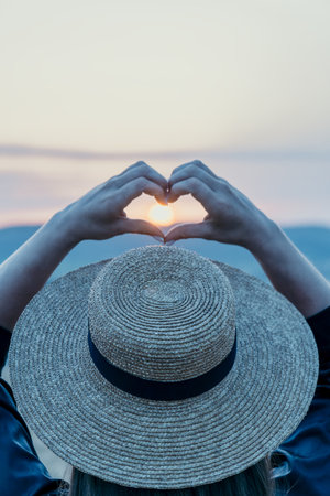 Sunset Heart Hands Hat: Woman forms heart shape sunset, peaceful moment, outdoor summer.の写真素材
