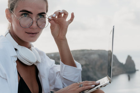 Woman Laptop Beach Work - Young woman with headphones and glasses works on laptop outdoors, looking...