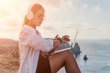 Woman Laptop Headphones Ocean - Young woman in white shirt working on laptop while listening to music with headphones on a cliff overlooking ocean during sunset.の写真素材