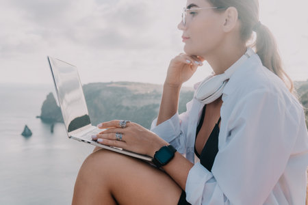 Woman Laptop Beach Working - A woman sits on a cliff overlooking the ocean, working on her laptop.