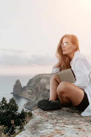 Woman, Cliff, Sunset: A young woman with glasses sits on a cliff overlooking the ocean at sunset, holding a notebook.の写真素材