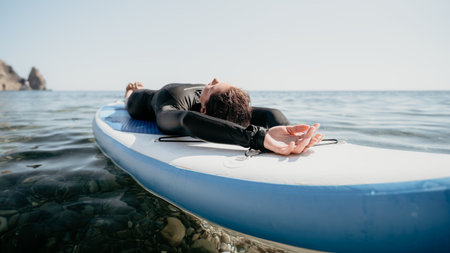 A person does yoga on a paddleboard in the ocean near a rocky cliff.の写真素材