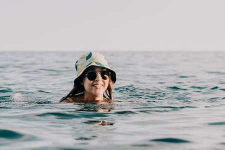 Woman Sunglasses Ocean - Smiling woman wearing sunglasses and a bucket hat swims in the ocean on a sunny day.の写真素材