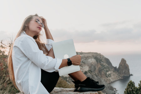 Woman Laptop Cliffside - Young woman working on laptop while sitting on a cliff overlooking the ocean at sunset.の写真素材