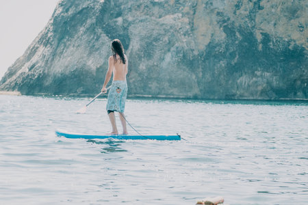 Man Paddleboarding Ocean Cliffside - A man paddles on a surfboard in the ocean near a cliffside.