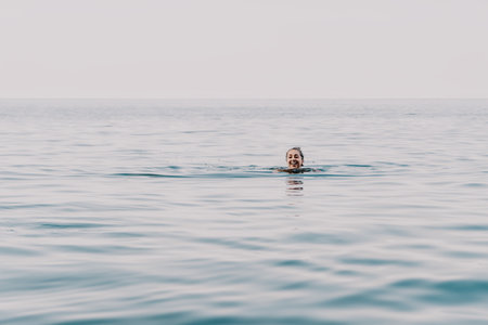 A swimmer is seen in the distance, a dark silhouette against the vast expanse of the ocean, as the sun dips below the horizon and the sky turns a soft orange.の写真素材