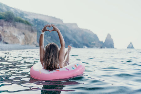 Woman, sea, relaxation. Young woman floats on pink donut in ocean making heart hands, enjoying...