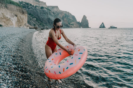 Woman, beach, ocean. Young woman in red swimsuit with donut inflatable entering water at pebble...