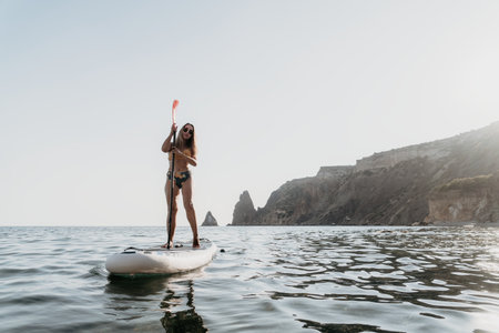 Woman Paddleboarding Ocean Coastline - A woman enjoys paddleboarding in the ocean off a rocky...