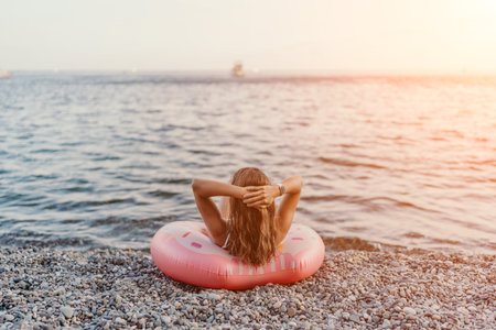 Woman beach ocean, relaxing on a pink donut inflatable ring at a pebble beach, enjoying a peaceful summer sunset vacation.の写真素材
