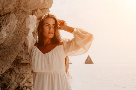 Woman White Dress Beach Seascape - A woman in a white dress stands by a rock formation on a beach with a boat sailing in the distance, enjoying the summer sun.の写真素材
